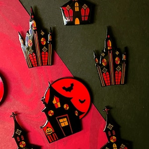 Könnte beinhalten: Eine Sammlung von Halloween-Anstecknadeln mit schwarzen Geisterhäusern und Kirchen mit roten und orangen Akzenten. Eine Nadel zeigt ein Haus vor einem roten Mond mit Fledermäusen. Die Anstecknadeln sind auf rotem und schwarzem Hintergrund angeordnet.