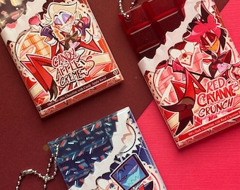 May include: Three rectangular chocolate bar keychains with ball chains. One features a blonde character and reads Candy Apple Creme, one features a red character and reads Red Cayenne Crunch, and one features a blue character and reads Blue Raspberry Byte.