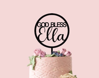 Op de afbeelding: Een tweelaagse taart versierd met roze en paarse frosting en bloemen. Een zwarte taarttopper met de tekst "God Bless Ella" in een cirkel zit bovenop de taart.