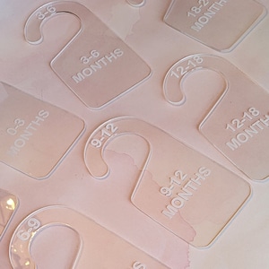 Puede incluir: Un conjunto de perchas de plástico transparente con texto blanco que indica diferentes rangos de edad para la ropa de bebé. Las perchas están etiquetadas con los siguientes tamaños: 0-3 meses, 3-6 meses, 6-9 meses, 9-12 meses, 12-18 meses y 18-24 meses.