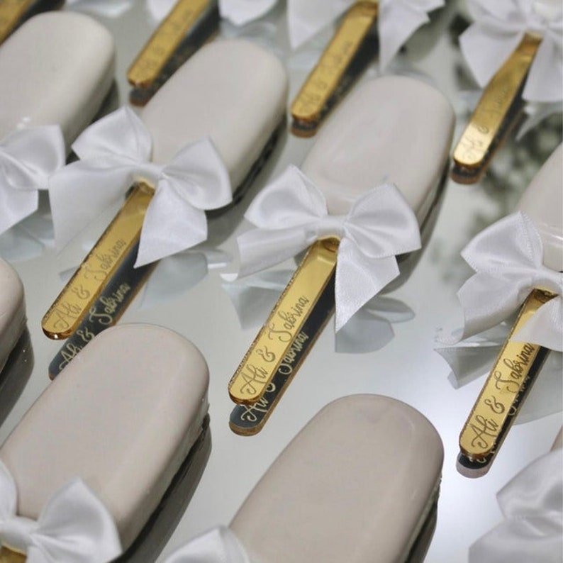 May include: White chocolate covered popsicle sticks with gold personalized sticks and white satin bows. The sticks are engraved with the names "Ali & Sakina" and the date "27.09.2022".