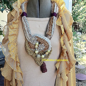 May include: A crocheted infinity scarf with a brown, beige, and gray color scheme. The scarf is decorated with beads and flowers. The scarf is displayed on a mannequin wearing a beige tank top and a gold lace shawl.
