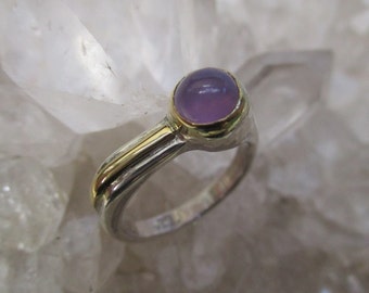 May include: A silver ring with a gold band and a round, lavender-colored stone. The ring has a smooth, polished finish and a simple, elegant design. The stone is set in a bezel setting, and the gold band adds a touch of warmth to the cool tones of the silver and lavender.