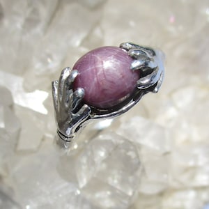May include: A silver ring with two hands holding a pink star ruby gemstone.