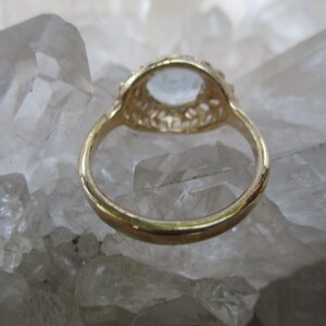 May include: A gold ring with a delicate filigree design and a clear gemstone in the center. The ring is set on a bed of white crystals.