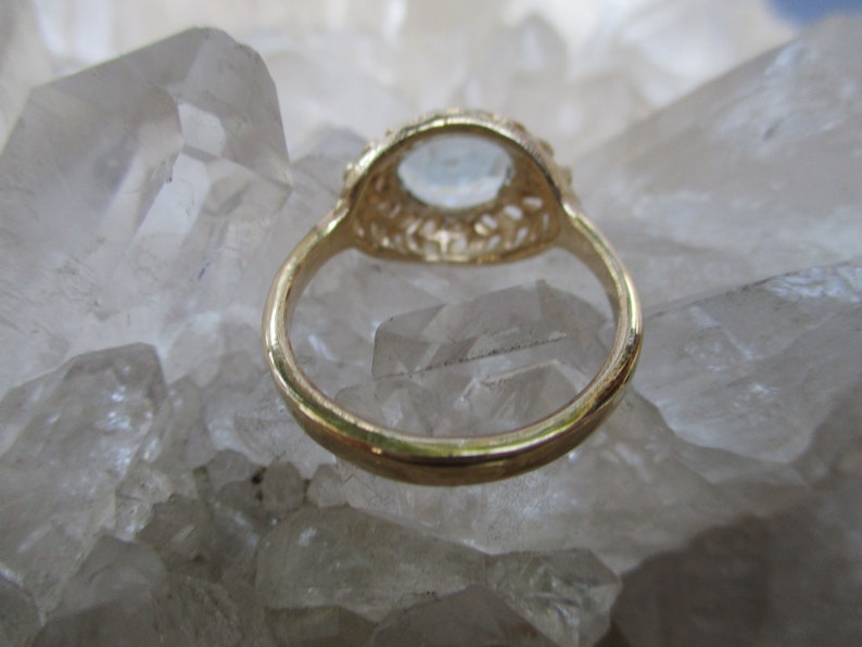 May include: A gold ring with a delicate filigree design and a clear gemstone in the center. The ring is set on a bed of white crystals.