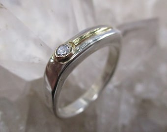 May include: A silver and gold ring with a single, clear diamond set in the center. The ring has a unique, curved design.