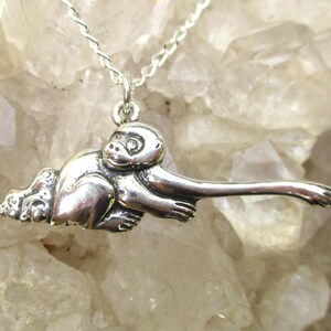 May include: A silver monkey pendant on a silver chain. The monkey is detailed with a long arm and outstretched hand. The pendant is suspended against a light-colored, textured background.