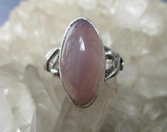 May include: A silver ring featuring a large, oval, light pink gemstone. The gemstone is set in a silver bezel. The ring's band has a decorative design with leaves and small beads. The ring is displayed on a white, textured surface.
