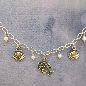 May include: A silver chain necklace with a mermaid charm and three shell charms. The necklace also has small white pearl beads hanging from it.