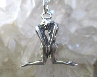 May include: A silver-toned metal pendant shaped like a figure kneeling with arms positioned behind the back. The pendant features a polished finish with dark recessed areas to define the contours of the body and is attached to a silver chain.