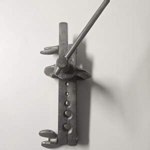 May include: A vintage metal flaring tool, featuring a handle and various sized holes. The tool is made of a dark, aged metal and is designed for shaping or expanding the ends of tubing.