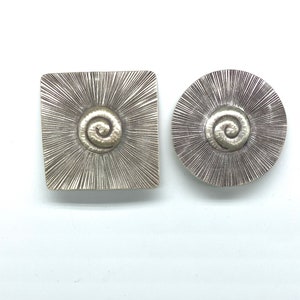 May include: Two silver metal decorative pieces with a textured surface and a spiral design in the center. One piece is square and the other is round.