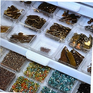 May include: A clear plastic storage box with multiple compartments filled with various small metal charms, beads, and findings. The box is organized with different types of embellishments, including hearts, stars, and geometric shapes.
