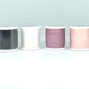 May include: Four spools of thread in a row, each with a different color. The colors are black, white, mauve, and light pink. The spools are on a white surface and have white bases and tops.