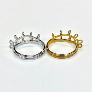 May include: Two adjustable rings, one silver-colored and one gold-colored, are displayed. Each ring features a decorative crown-like design with multiple prongs. The rings are set against a plain white background.
