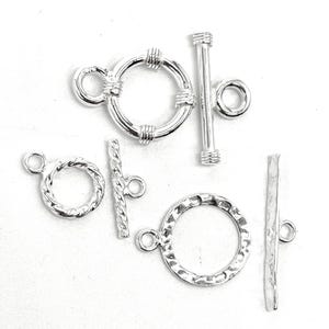 May include: Two silver-toned toggle clasps for jewelry making. One clasp features a smooth ring with ribbed accents and a plain bar. The second clasp consists of a twisted rope-textured ring and a matching twisted bar, both in a metallic finish.