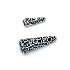 May include: Two silver cone-shaped bead caps with a decorative pattern of small circles. The caps are made of metal and have a shiny finish.