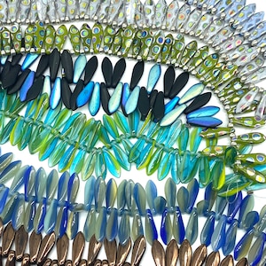 May include: Assortment of glass beads in various colors and patterns, including blue, green, brown, and iridescent. The beads are shaped like teardrops and are arranged in rows.