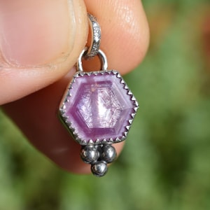 May include: A hexagonal pink gemstone pendant set in a silver bezel with a serrated edge. The pendant features a small silver bail at the top and a cluster of three small silver spheres attached to the bottom point of the hexagonal setting.