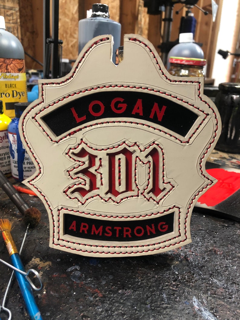 May include: White leather firefighter helmet patch with red stitching. The patch features the name "LOGAN" in black letters with red outlines, the number "301" in red with white outlines, and the last name "ARMSTRONG" in black letters with red outlines.