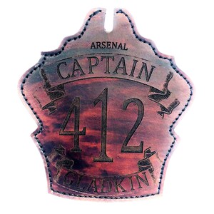 May include: A brown leather fire helmet patch with the text "ARSENAL CAPTAIN 412 GLADKIN" in black lettering.