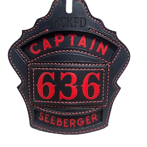 May include: Black leather firefighter helmet shield with red stitching. The shield has the text "KCKFD CAPTAIN 636 SEEBERGER" in red lettering.