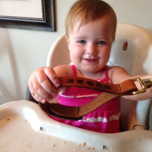 May include: A young child wearing a pink and white striped shirt is sitting in a high chair. The child is holding a brown leather strap with the word "BROWN" embossed on it.