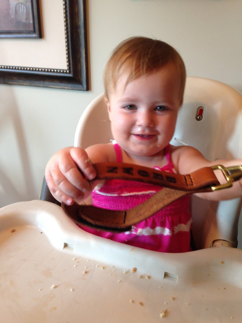 May include: A young child wearing a pink and white striped shirt is sitting in a high chair. The child is holding a brown leather strap with the word "BROWN" embossed on it.