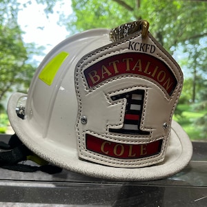 May include: White firefighter helmet with a leather patch that reads "KCKFD Battalion 1 Cole".