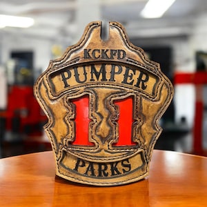 May include: A tan leather firefighter helmet shield with the text "KCKFD PUMPER 11 PARKS" in black. The number 11 is in red. The shield is sitting on a wooden surface.