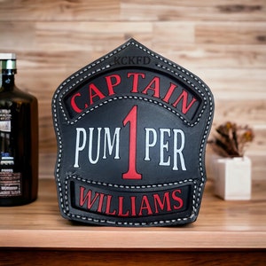 May include: Black leather firefighter helmet shield with white stitching. The shield has the text "KCKFD CAPTAIN PUM 1 PER WILLIAMS" in white and red lettering.