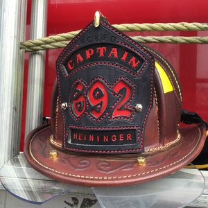 May include: A black leather firefighter helmet with a red and black shield. The shield has the text "KCFD CAPTAIN" and the number "692" in red. The helmet also has the name "HEININGER" in black.