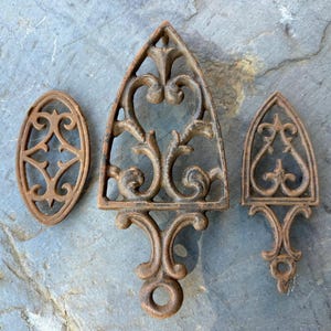 May include: Three antique cast iron trivets with intricate floral designs. The trivets are in various shapes, including a heart, an oval, and a pointed arch.