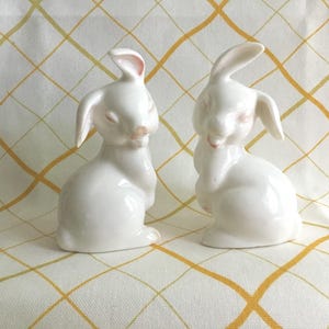 May include: Two white ceramic bunny figurines with pink accents on a yellow and white checkered tablecloth. The bunnies have long ears and are in a seated position, with their eyes closed.
