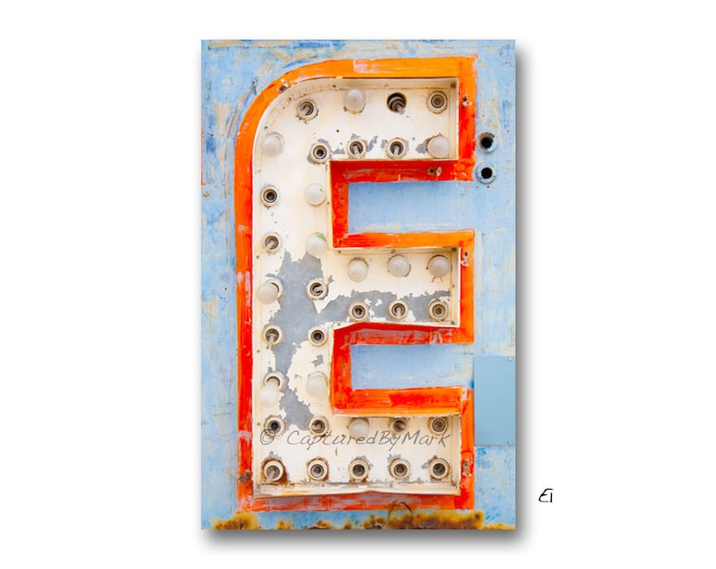 K&ouml;nnte beinhalten: Ein vintage, verwittertes, orange-wei&szlig;es Neon-Schild mit dem Buchstaben "E" in der Mitte. Das Schild hat einen blauen Hintergrund und ist leicht verblasst. Der Buchstabe "E" besteht aus Gl&uuml;hbirnen, die an einigen Stellen fehlen.