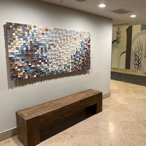 May include: A wooden bench with a dark brown finish sits in front of a wall with a large abstract art piece made of colorful squares. The art piece is a mosaic of blues, greens, browns, and whites.