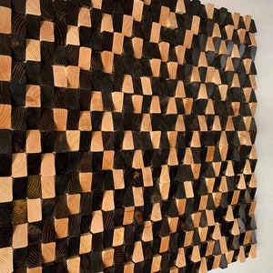 May include: A wall art piece made of wood blocks in a geometric pattern. The blocks are arranged in a staggered pattern, creating a 3D effect. The blocks are painted in black and natural wood tones.