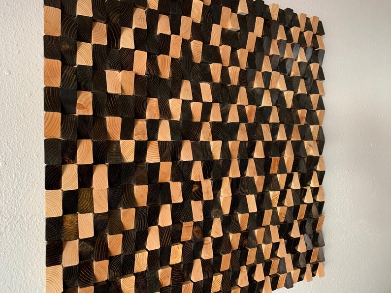 May include: A wall art piece made of wood blocks in a geometric pattern. The blocks are arranged in a staggered pattern, creating a 3D effect. The blocks are painted in black and natural wood tones.