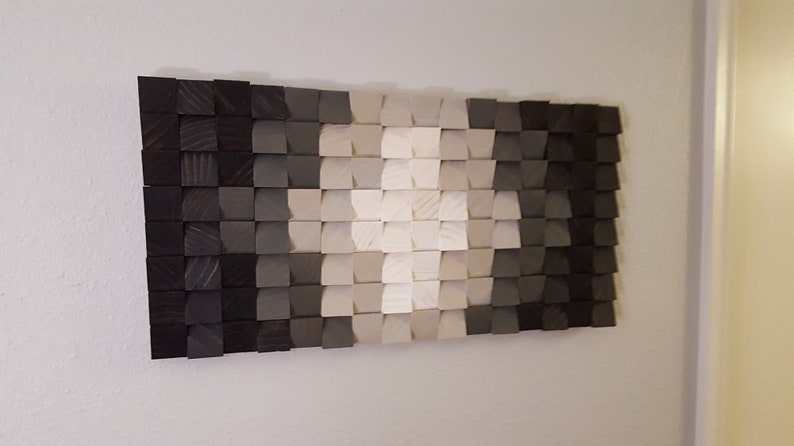 May include: A wall art piece made of wood blocks in shades of gray and white. The blocks are arranged in a gradient pattern, creating a modern and abstract design.