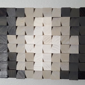 May include: A wall art piece made of wood blocks in various shades of gray and white. The blocks are arranged in a geometric pattern, creating a gradient effect from dark gray to white.