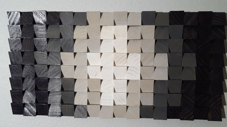 May include: A wall art piece made of wood blocks in various shades of gray and white. The blocks are arranged in a geometric pattern, creating a gradient effect from dark gray to white.