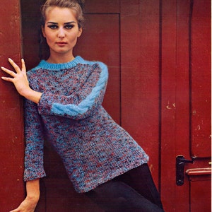 Pode incluir: Uma mulher vestindo um suéter azul e vermelho com padrão e mangas azuis. Ela está em pé em frente a uma porta vermelha.