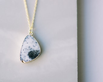 May include: A gold-toned necklace featuring a pendant with a white and black speckled stone. The stone has a gold-colored trim and is suspended from a delicate gold chain. The pendant has a unique, organic shape.