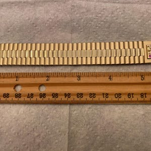 May include: A gold-toned metal watch band with a clasp that reads "PC&E". The band is approximately 5 inches long and has a textured surface.