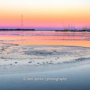 May include: A pink and orange sunset over a frozen lake with a small town in the distance. The lake is covered in ice and snow, and the sky is a beautiful blend of colors.