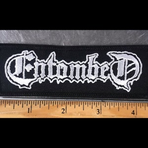 May include: White embroidered "Entombed" logo patch on a black fabric background. The patch is approximately 4 inches long.
