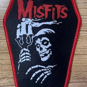 May include: Black and red embroidered patch with the word "Misfits" in red lettering and a white skeleton holding a trident. The patch is shaped like a coffin.