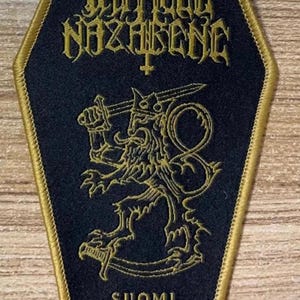May include: Black and gold embroidered patch shaped like a coffin with the text "DEFILED NAZARENE" and a lion with a sword. The text "SUOMI FINLAND PERKELE" is at the bottom of the patch.