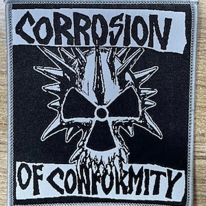 May include: Square black patch with the words "CORROSION" and "OF CONFORMITY" in white, stylized text. A white skull with a radiation symbol is in the center, surrounded by sharp spikes. The patch has a gray border.